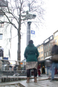 Eine Frau steht, mit dem Rücken zur Kamera, vor einem Mast mit vielen Überwachungskameras und schaut hinauf. Ihre Hände hat sie in die Jackentaschen gesteckt.